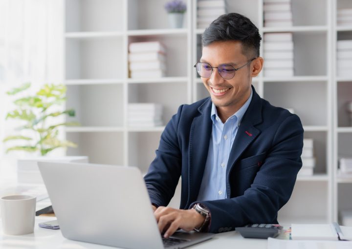 Man in glasses and business casual clothes smiling while typing on a laptop